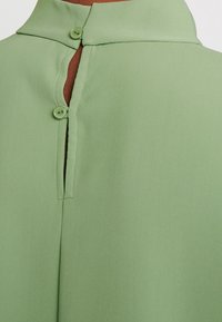 Blusa verde chiaro con colletto a giro, chiusura con bottoni sul retro e dettaglio a spacco, realizzata in un tessuto liscio con una texture sottile.