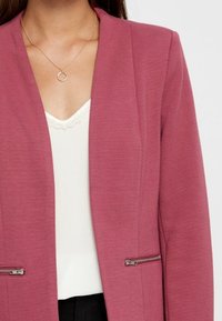 Růžový blazer s texturovanou tkaninou, se dvěma zipovými kapsami, nošený přes bílý top s krajkovým detailem na výstřihu.