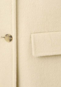 Priblížený záber na béžový vlnený kabát so zapínaním na gombík vľavo a obdĺžnikovým náprsníkovým vreckom vpravo.