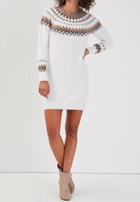 Femme portant une robe pull en maille blanche avec des motifs géométriques marron et noirs, ainsi que des bottines beige, debout avec une main sur la hanche.