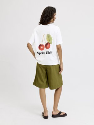 Person med krøllete hår står med ryggen til, iført en hvit T-skjorte med kirsebær og teksten "Spring Vibes", olivengrønne shorts og svarte sandaler.