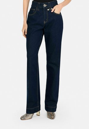 Dark denim wide-leg jeans with visible stitching, high waist, front pockets, and a metallic button closure. Styled with patterned ankle boots.