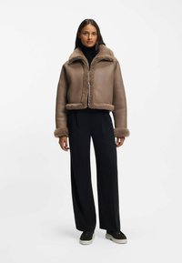 Brązowa skórzana kurtka z miękkim futrzanym podszyciem, krótkim fasonem, suwakiem z przodu i mankietami obszytymi futrem, zestawiona z ciemnymi, szerokimi spodniami.