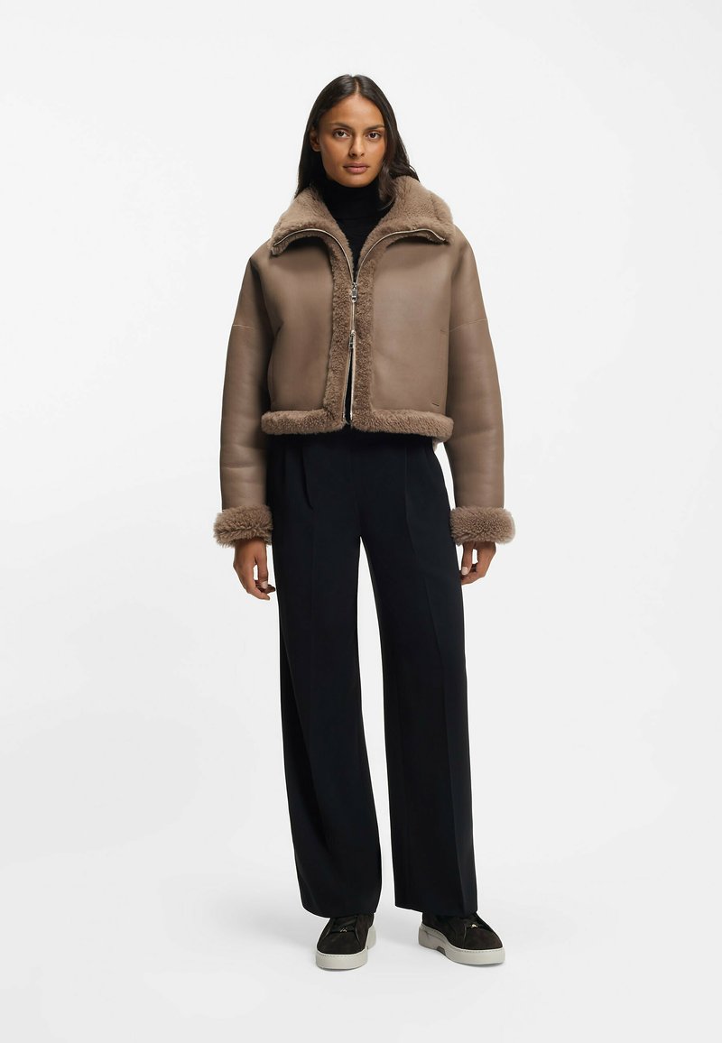 Brązowa skórzana kurtka z miękkim futrzanym podszyciem, krótkim fasonem, suwakiem z przodu i mankietami obszytymi futrem, zestawiona z ciemnymi, szerokimi spodniami.