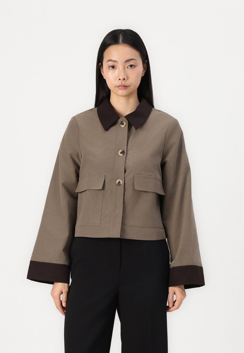 Brun cropped jakke med en mørk krage, to frontlommer og knappestenging. Har lange ermer med en mørkere mansjett detalj.