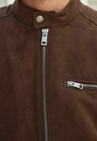 Giacca in suede marrone con chiusura frontale a cerniera, colletto con bottone a pressione e tasca sul petto con cerniera orizzontale.