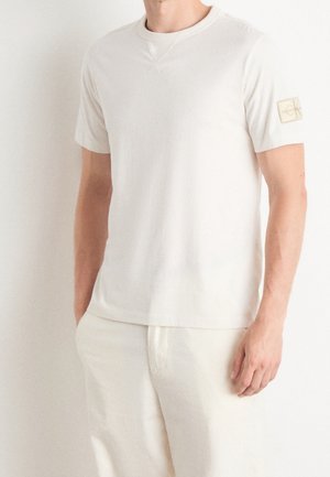Mann, der ein schlichtes weißes T-Shirt mit kurzen Ärmeln und weiße Hose trägt, steht mit einer Hand in der Tasche vor einem weißen Hintergrund.