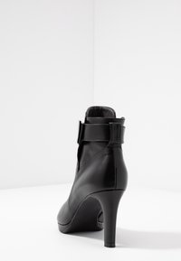 Černá kožená vysoká kotníková bota na jehlovém podpatku s elegantním designem, která má výrazný nastavitelný pásek a hladkou texturu.
