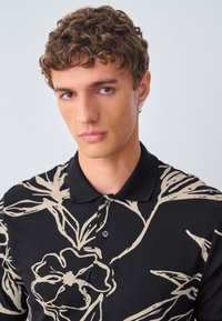 Juodas polo marškinėlis su dideliu, abstrakčiu smėlinės spalvos gėlių raštu, su apykakle ir trimis mygtukais priekyje. Lygus audinio paviršius.