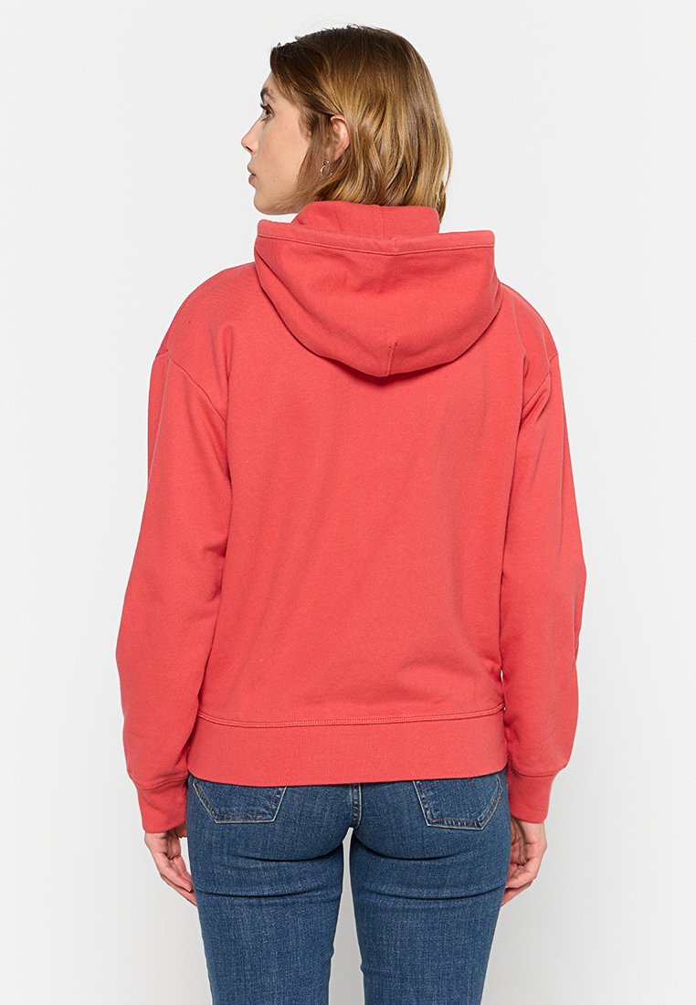 Felpa rossa con cappuccio regolabile, polsini e bordo a costine. Il tessuto sembra morbido. Indossata con jeans in denim blu. Vista posteriore semplice.