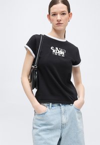Νεαρή γυναίκα που φοράει μαύρο t-shirt Calvin Klein 1968, ανοιχτό μπλε τζιν και μαύρη τσάντα ώμου, στέκεται με τα χέρια στις τσέπες.