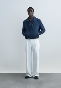 Moški stoji ob ravni steni, oblečen v mornarsko modro jakno na zadrgo, modro srajco, bele široke hlače in črne čevlje, z rokami v žepih jakne.