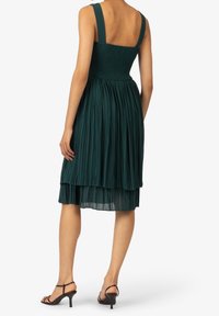 Grön plisserad klänning med smockad byst och breda axelband, med en lager på lager nederdel. Bärs med svarta klacksandaler.