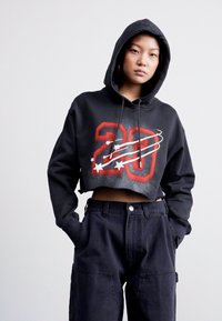 Black cropped hoodie with a red graphic "20" and white star accents, featuring a drawstring hood and soft material. Worn with high-waisted dark pants.