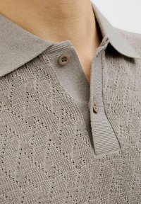 Vaaleanharmaa neulepolo, jossa on tekstuurinen aaltoilukuosi. Pololla on pehmeä kaulus ja kaksi ruskeaa nappia kaula-aukossa.