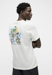 Άνδρας με σταυρωμένα χέρια, που φοράει λευκό t-shirt The North Face με πολύχρωμο αφηρημένο κείμενο και λουλουδάτο σχέδιο στην πλάτη.