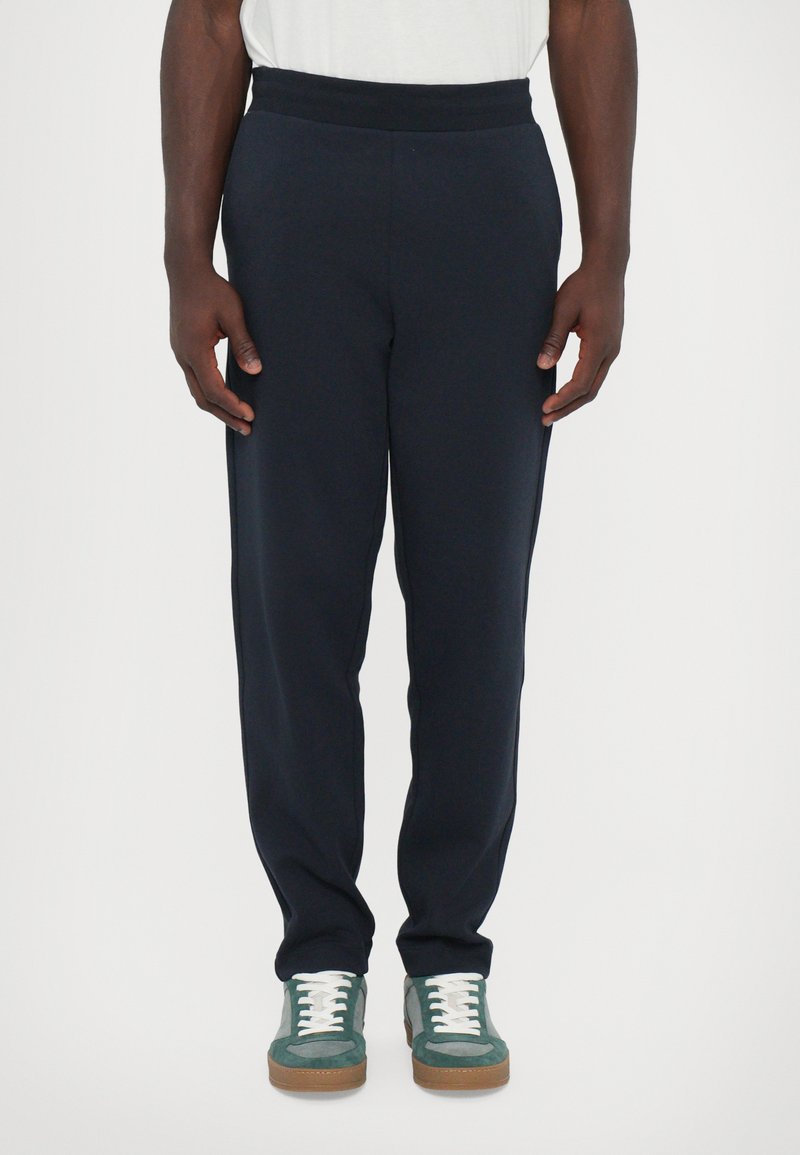 Navy blue sweatpants with elastic waistband, tapered legs, side pockets, paired with green and white sneakers with brown soles.