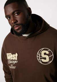 Felpa marrone con ricamo "West Chicago" e una toppa a tema basket con una "S". Tessuto morbido e vestibilità comoda.