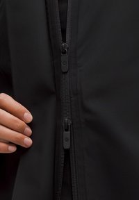Ruka drži djelomično raspovijen dvostruki patentni zatvarač na crnoj jakni, prikazujući detalje zatvarača i teksturu tkanine.