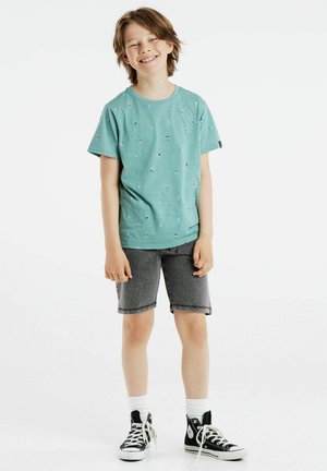 Groen T-shirt met korte mouwen met gekleurde kleine patronen, gecombineerd met grijze denim shorts. Zwarte hoge sneakers en witte sokken zichtbaar.