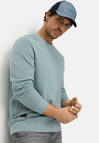 Lichtblauwe gebreide trui met ronde hals en geribbelde manchetten. Draagt een grijze spijkerbroek en een pet met een lichtblauw en marineblauw ontwerp.