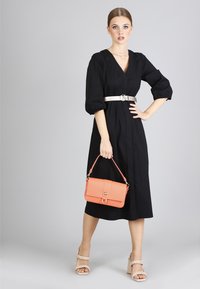 Abito midi nero con maniche a palloncino, cintura argentata e borsa in coral textured. Modella indossa sandali con tacco a blocco beige. Sfondo semplice.