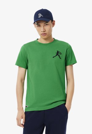 Groene katoenen T-shirt met een ronde hals en korte mouwen, voorzien van een zwarte afbeelding van een rennende figuur op de borst. Draagt onder een marineblauwe pet.
