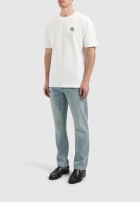 Witte katoenen t-shirt met een klein zwart logo, gecombineerd met lichtblauwe denim jeans en zwarte leren schoenen, staande op een neutrale achtergrond.
