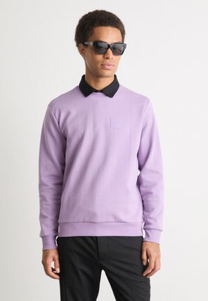 Felpa viola a maniche lunghe con colletto nero, polsini a costine e un patch sul petto. Il modello indossa occhiali da sole neri e pantaloni neri.