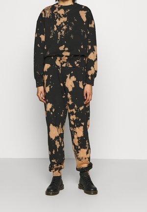 Personne portant un sweat-shirt noir et marron éclaboussé d'eau de Javel et un pantalon de jogging assorti avec des bottes noires à lacets sur fond clair.