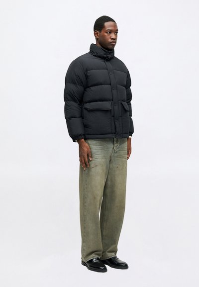 Vyras stovi vilkėdamas juodą pūkinę striukę, laisvus išblukusius žalius džinsus su dėmėmis ir juodus odinius batus prieš baltą fono.