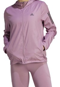 Helle rosa Sportjacke mit Kapuze, Reißverschluss und Fronttaschen. Mit schwarzen Akzenten in drei Streifen an den Ärmeln und einem Logo auf der Brust.