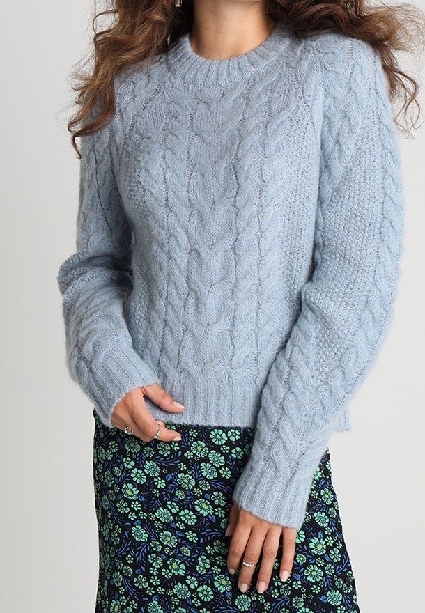 Hellblauer Kabelstrickpullover mit rundem Ausschnitt und gerippten Bündchen, kombiniert mit einem Röcken mit Blumenmuster in Grün- und Marineblau-Tönen.