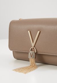 Ruskea teksturoidusta nahasta valmistettu laukku, jossa on kultasävyinen V-muotoinen lukko ja koristeellinen ketjuhapsu. Kompakti suorakaiteen muotoinen malli siisteillä linjoilla.