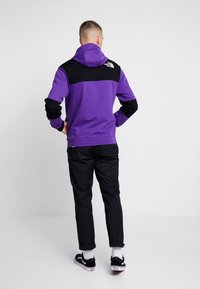 Lila hoodie med svarta detaljer på axlar och ärmar, texturerat tyg, känguruficka, matchad med svarta byxor och svarta sneakers.