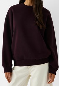 Mujer con un sudadera oversized oscura de color púrpura y mangas largas, junto con pantalones blancos, frente a un fondo liso.