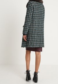 Vävd kappa i blå och grön rutigt mönster med pälskrage, med långa ärmar och rak skärning, kombinerad med en mörk läderkjol och ankelstövlar.