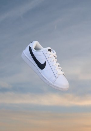 Baskets blanches Nike avec logo swoosh noir flottant sur un fond de ciel bleu et orange.