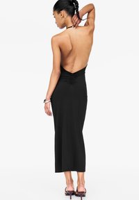 Robe noire sans manches avec un dos en V profond, détail froncé à la taille, et une jupe longueur sol, accompagnée de chaussures à talons hauts.