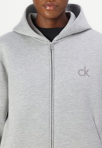 Šedá mikina so zipsom a dvojito podšívkovanou kapucňou, s malým vyšitým logom "ck" na ľavej strane hrudníka a hladkou, mäkkou textúrou.