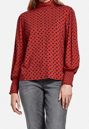 Vrouw draagt een rood gebloemde blouse met lange mouwen, met gerimpelde manchetten en kraag, gecombineerd met vervaagde grijze jeans.