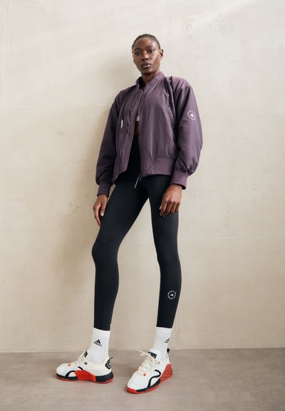 Lila bombázódzseki, fekete leggings és fehér sportcipő piros részletekkel; rétegezett textúrák és sportos dizájn elemek.