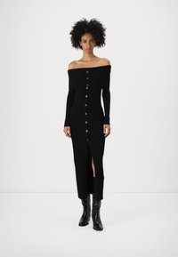 Svart ribbat off-shoulder klänning med knäppning framtill, långa ärmar och hög slits, matchad med glänsande svarta ankelstövlar.