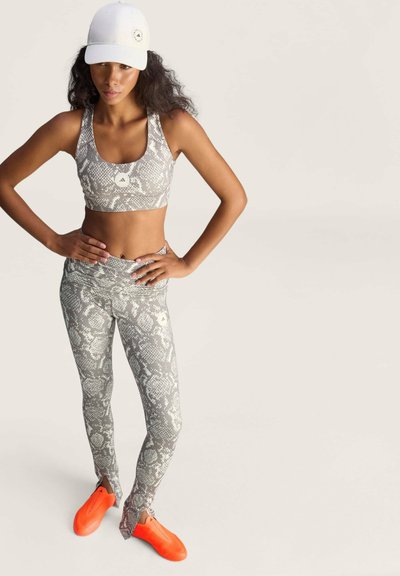 Vrouw draagt een grijze sport-bh en legging met slangenprint, witte pet en feloranje slip-on schoenen, staat met handen in de heupen.