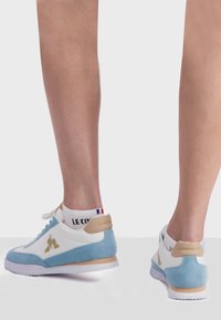 Sneakers bianchi e azzurri chiaro con materiali in suede e pelle. Presentano una suola testurizzata, lacci abbinati e un logo dorato sul lato.