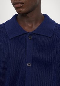 Navy blue knitted cardigan with a textured surface, featuring a classic collar and two black buttons down the front.
