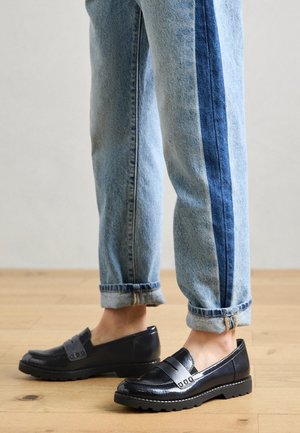 Ben iført opbukket tofarget blå jeans og sorte læderloafers, stående på et trægulv.
