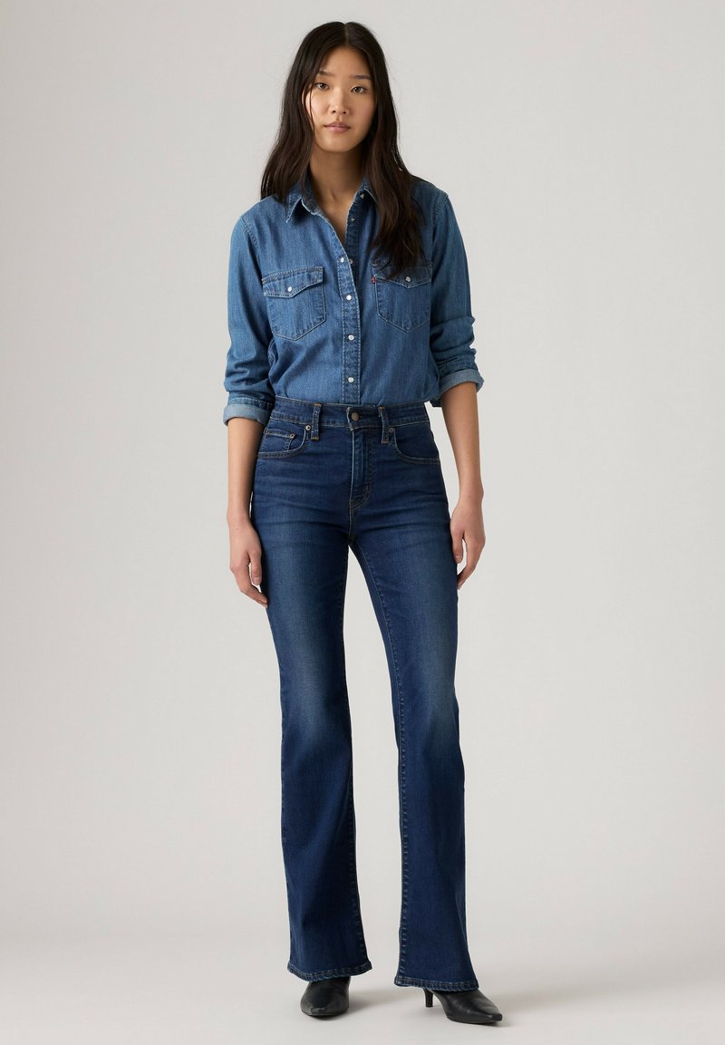Femme aux longs cheveux foncés portant une chemise en jean bleue et un jean flare taille haute bleu foncé avec des chaussures à talons noires.
