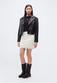 Μαύρο cropped δερμάτινο μπουφάν biker, μαύρο t-shirt με λογότυπο CK, λευκή υφασμάτινη φούστα ψηλόμεση και μαύρες μπότες μέχρι το γόνατο.
