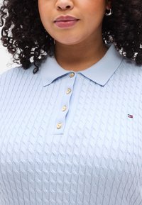 Ανοιχτό γαλάζιο πλεκτό πόλο με υφαντό διακοσμητικό μοτίβο σε σχήμα ρόμβου, με γιακά και πέντε χρυσά κουμπιά. Μικρό λογότυπο στην αριστερή πλευρά.